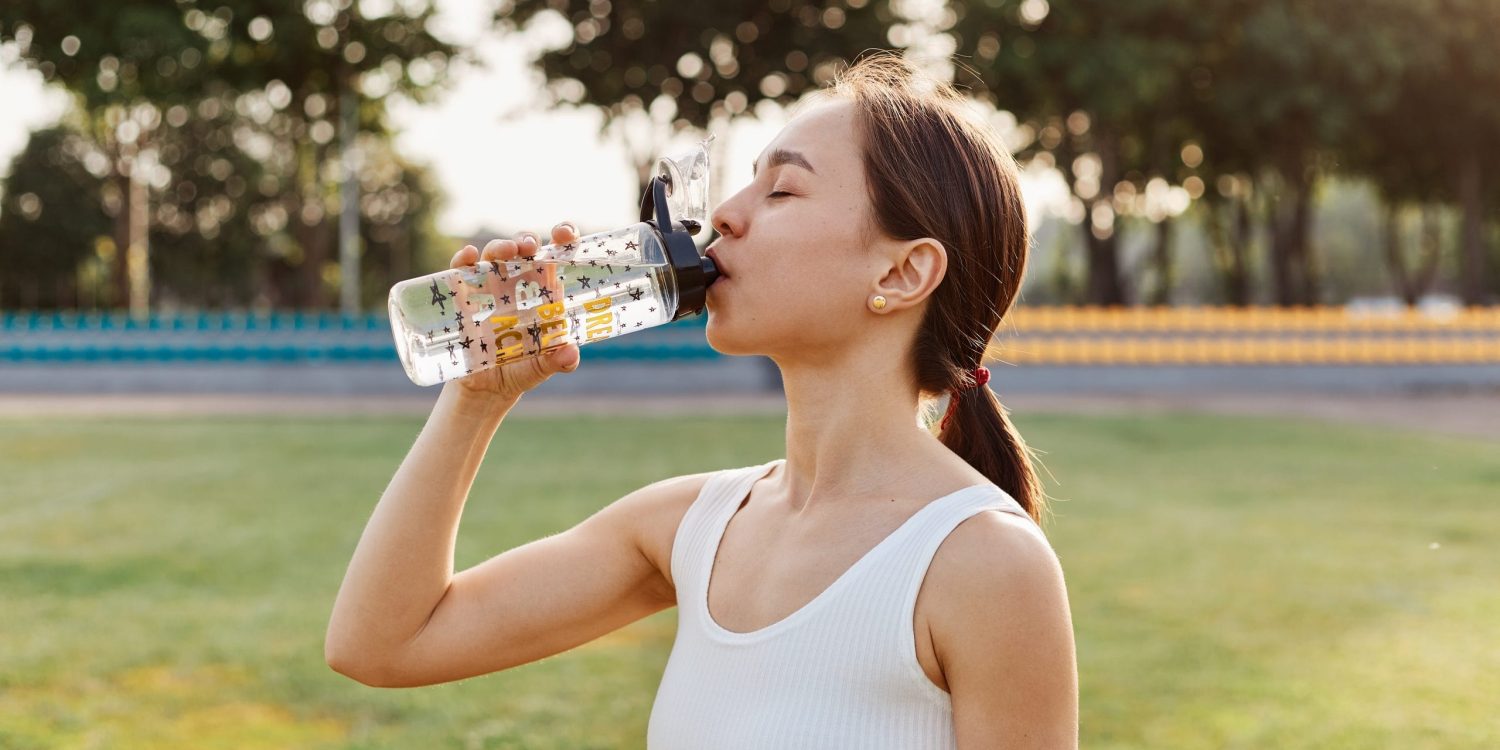 Fit dark haired female drinking water from her water bottle on stadium just before she starts training, outdoor workout in summertime, healthy lifestyle. kobieta pije wode nawodnienie